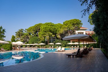 Boş güverte sandalyeleri ve şemsiyeler, yemyeşil ağaçlı yüzme havuzu ve turizm merkezinin arka planında açık mavi gökyüzü tarafından düzenlenir.