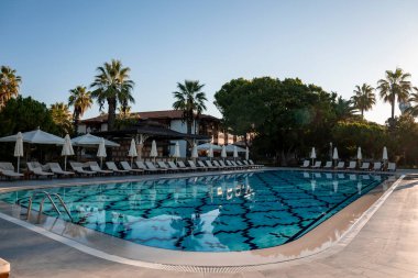 Boş şezlonglar ve katlanır şemsiyeler saydam yüzme havuzunun etrafına yerleştirilmiş ve turist otelinin arka planında açık gökyüzü var.