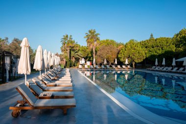 Boş şezlonglar ve katlanır şemsiyeler yüzme havuzunun etrafına dizilmiş ağaçlar ve turist otelinin arka planında açık mavi gökyüzü.