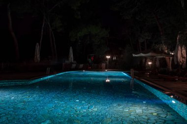 Şemsiyeler ve boş şezlonglar aydınlatılmış yüzme havuzunun yanında. Geceleri arka planda ağaçlar var.