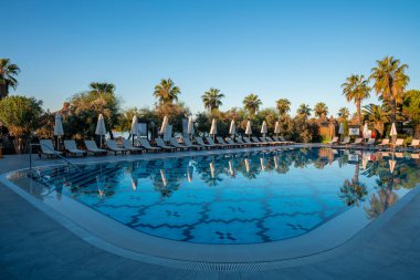 Boş güverte sandalyeleri ve katlanır şemsiyelerle çevrili havuz kenarında sıralanmış ağaçlarla turizm merkezinin arka planında açık mavi gökyüzü