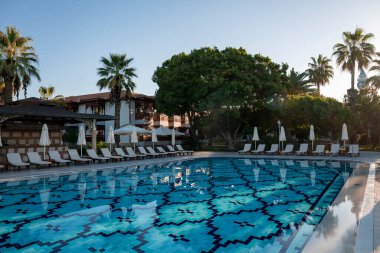 Boş şezlonglar ve katlanmış şemsiyeler şeffaf yüzme havuzunun etrafına yerleştirilmiş ve turizm merkezinin arka planında açık gökyüzü var.