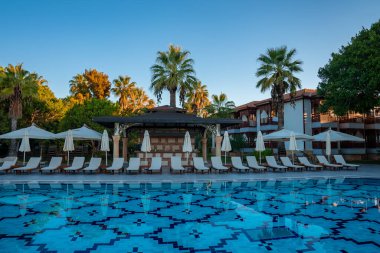 Boş şezlonglar ve katlanmış şemsiyeler turistik bir tatil beldesinde, arka planda palmiye ağaçları ve açık mavi gökyüzü ile düzenlenmiştir.