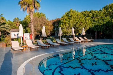 Boş güverte sandalyeleri ve katlanır şemsiyelerle çevrili havuz kenarında sıralanmış ağaçlarla turizm merkezinin arka planında açık mavi gökyüzü