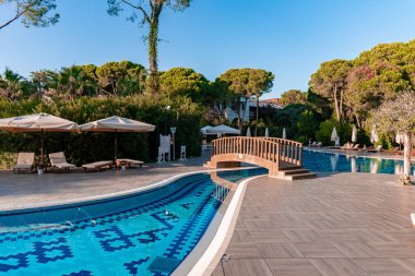 Güneşli bir günde turist merkezinde açık mavi gökyüzünün altında yemyeşil ağaçlarla çevrili yüzme havuzu üzerindeki yaya köprüsü manzarası