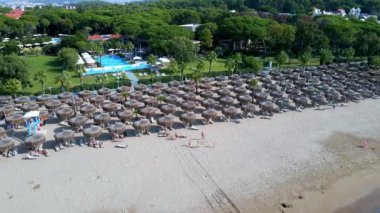 Şemsiyelerin havadan görünüşü, kumlu sahilde palmiyeler, insanlar, gün batımında dalgalı mavi deniz. Yaz tatili. Palmiye ağaçları, şemsiyeler, beyaz kum ve okyanusla tropik bir manzara..