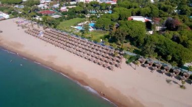 Şemsiyelerin havadan görünüşü, kumlu sahilde palmiyeler, insanlar, gün batımında dalgalı mavi deniz. Yaz tatili. Palmiye ağaçları, şemsiyeler, beyaz kum ve okyanusla tropik bir manzara..