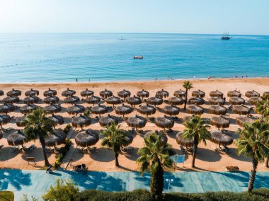 Şemsiyelerin havadan görünüşü, kumlu sahilde palmiyeler, insanlar, gün batımında dalgalı mavi deniz. Yaz tatili. Palmiye ağaçları, şemsiyeler, beyaz kum ve okyanusla tropik bir manzara..