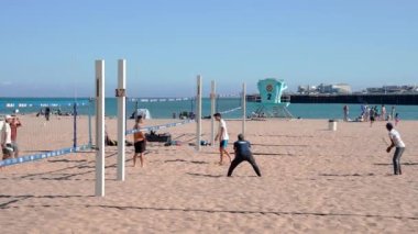 People playing volleyball at sandy beach with Santa Cruz Municipal Wharf in the background under clear blue sky during sunny day
