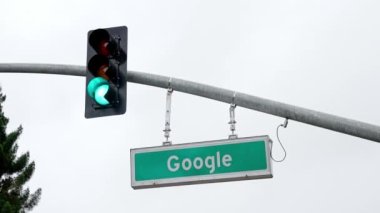 Google text on sign board hanging by traffic signal on pole below cloudy sky. Google street sign.