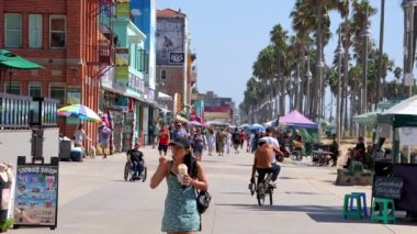 Tourists walking on boardwalk by palm trees and exploring shops at the Venice beach on a warm sunny day