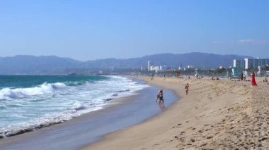 Waves splashing on shore and people enjoying sandy beach in California. Palm trees growing by the Pacific ocean. Sunny day in Los Angeles by the beach.