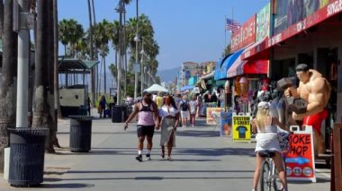 Tourists walking on boardwalk by palm trees and exploring shops at the Venice beach on a warm sunny day