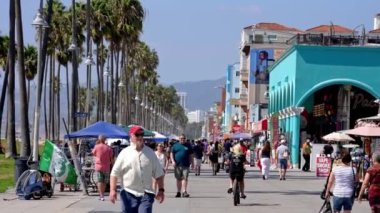 Tourists walking on boardwalk by palm trees and exploring shops at the Venice beach on a warm sunny day