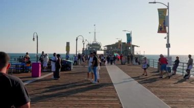 Ferris wheel and roller coaster rides and people exploring shops at the Pacific amusement park on the Santa Monica Pier.