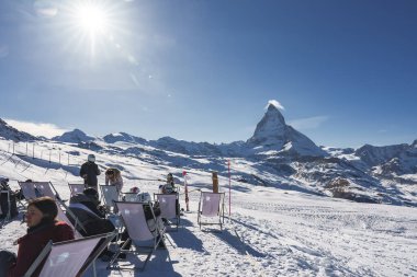 Erkek ve kadın turistler, kar üzerinde iskemlede dinleniyorlar. Kayak merkezinin parlak güneşli manzarası ve İsviçre 'nin Zermatt şehrinin arka planında açık mavi gökyüzü var.