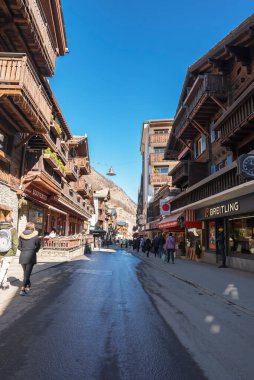 Erkek ve kadın turistler, İsviçre 'nin Zermatt şehrinde arka planda açık mavi gökyüzü ve lüks otel binaları arasında sokakta yürüyorlar.