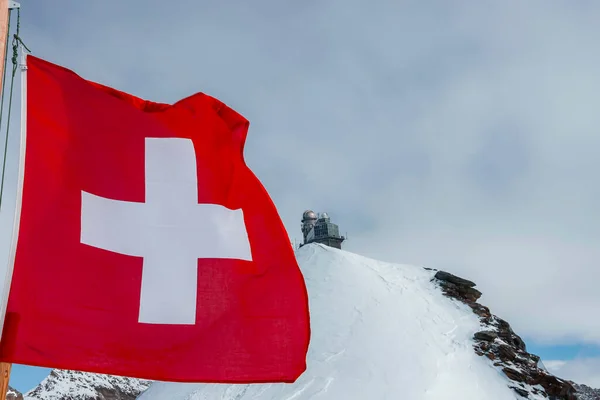 Swiss flag waving on pole with view of Jungfrau ice palace on top of snow covered mountain under cloudy sky at Switzerland, winter holiday travel concept