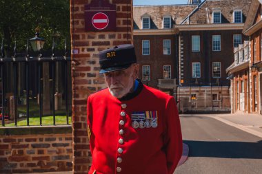 Yaşlı emekli, Royal Chelsea hastanesinin önünde duran kırmızı üniformalı. Eski asker, arka plandaki huzurevini düşünüyor. Güneşli bir günde dinleniyor..