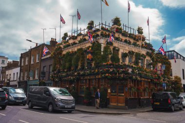 Churchill Arms çiçek ve yapraklarla süslenmiş. Arabalar şehirde yolda ilerliyor. Londra 'nın ünlü barının önünden geçen bir adam..