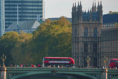 Westminster Köprüsü 'nde ağaçlar arasında turnuva otobüsünün yanından geçen kalabalığın uzaktan görüntüsü. Turistler saray mimari yapıyı keşfediyorlar. Arka planda gökyüzü olan modern ve antik binaların manzarası.