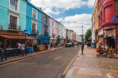 Portobello Yolu 'ndaki Notting Hill' den alışveriş yapan turistler. Moda, antika ve eski moda satan işlek pazarı ile ünlüdür. Arka planda bulutlu gökyüzü olan kafeler ve dükkanlar.