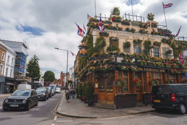 Churchill Kolları çiçek ve yapraklarla kaplı. Arabalar bulutlu gökyüzünün altında yolda ilerliyorlar. İnsanlar Londra 'nın ünlü barının önünden yürüyorlar..