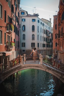 A romantic couple stands on a bridge in Venice, overlooking a narrow canal and surrounded by charming buildings and alleyways, creating a picturesque and serene atmosphere. clipart