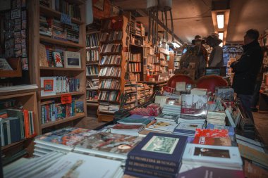 Rafları kitaplarla dolu loş bir kitapçı. Oturma alanı ya da dekoratif öğeler yok. Özel bölümler, kitap türleri ve mimari tarzı bilinmiyor.