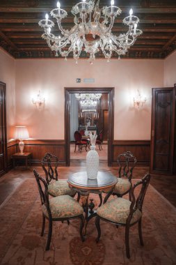 Avizesi ve ahşap masası olan lüks bir oda. Bilinmeyen açı ve konum. Ahşap döşeme. Tarz ve materyal belirtilmemiş. Görünür sanat eseri ya da perde yok..