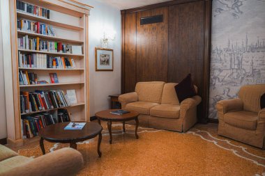 Koltuğu, sehpası ve kitaplığı olan rahat bir dairede iyi ışıklandırılmış, kahverengi ve bej renkli bir oturma odası. Doğal ışık ve tenha aydınlatma sıcak ve davetkar bir atmosfer yaratır..