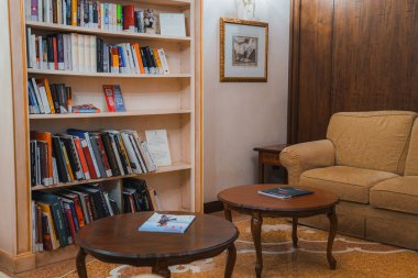 Cozy living room in Venice, Italy with a brown couch, coffee table, and bookcase. Neutral color scheme and no windows. Part of a collection showcasing Venetian hotel and restaurant interiors. clipart