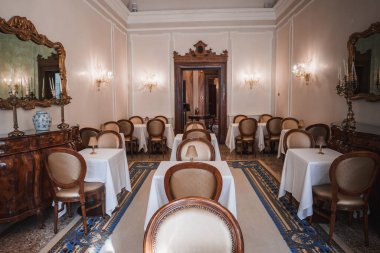 Masaları ve sandalyeleri olan şık ve süslü bir yemek odası. Dekor bilinmeyen bir dönemden esinlenilmiştir. Belirli bir işaret ya da aydınlatma armatürü yoktur..