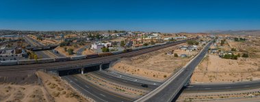 Yeşil alanlar, yeşil alanlar ve açık mavi gökyüzünün altındaki uzak dağlarla bej binalar ve tepe kenti boyunca uzanan eğimli bir otoyolun havadan görüntüsü..