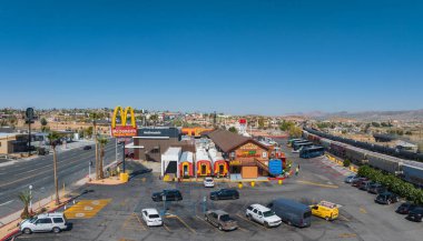 McDonalds gibi ikonik yiyeceklerle dolu hareketli bir Barstow ticaret merkezinin havadan görünüşü, bir dağ zeminine, berrak gökyüzüne ve geçen bir trene karşı kurulmuştur..