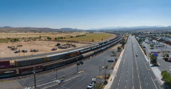 Aerial view of a serene semi-arid landscape with mountains, a road with parked vehicles, a moving freight train, and a modern town under a clear blue sky.