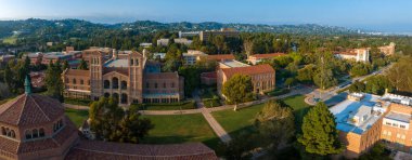 UCLA kampüsünün havadan görünüşü Gotik kulesi, kırmızı tuğlalı binalar, yeşil çimenler ve tepecikli ağaçlarla kaplı yumuşak, altın renkli manzaralı yollar..