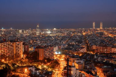Barcelona, İspanya 'nın panoramik gece manzarası, parlayan pencereleri, merkezi ışıklı bir yolu, ve Torre Glories ve Hotel Arts gibi simgeleri alacakaranlık gökyüzü altında.
