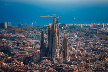 Barcelona 'nın panoramik manzarasında Sagrada Familia' nın kule kuleleri ve vinçleri var. Şehir Akdeniz mimarisi ve açık bir gökyüzünün altındaki mavi deniz ufku görülebilir..
