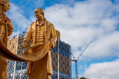 Birmingham, İngiltere 'de bulunan iki tarihi şahsın altın heykeli Birmingham Kütüphanesi' nin bal peteği cephesiyle ve parçalı bulutlu bir gökyüzünün altında bir inşaat vinciyle çelişiyor..