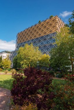 Birmingham, İngiltere 'deki modern bir bina, açık mavi gökyüzüne karşı gün ışığında parlayan bir bal peteği cephesiyle övünüyor. Canlı yeşilliklere sahip yemyeşil bahçeler doğal bir kontrastlık sağlar..