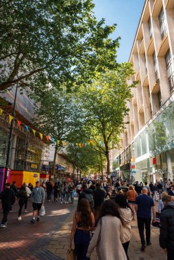 Birmingham, İngiltere 'de güneşli bir günde modern binalar, yeşil gölgeler ve benekli ışıklarla dolu canlı bir cadde sergileniyor. İnsanlar renkli kiraz kuşları altında gezer, canlı şehir atmosferinin tadını çıkarırlar..