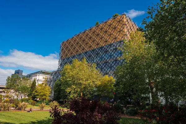 Birmingham, İngiltere 'de mavi gökyüzüne karşı parıldayan, yeşil ve çiçek açan kırmızı çiçeklerle çevrili, şehrin mimari füzyonunu gözler önüne seren, bal peteği görünümlü modern bir bina..