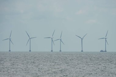 Büyük rüzgâr türbinleri bulutlu bir gökyüzünün altında Kopenhag ve Malmö arasındaki ufka doğru ilerlerken, ölçeklendirmek için son türbine yakın bir botla sakin bir denizde dönüyor..