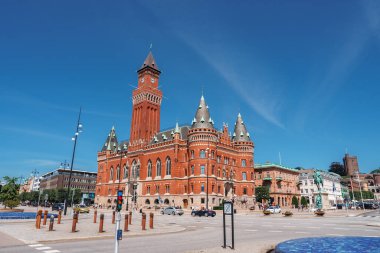 Romanesque ve Gotik Diriliş elementlerine sahip, içinde kule ve kemerli pencereler bulunan güneş ışığında kırmızı tuğlalı bir bina, Helsingborg 'da sessiz bir plazada duruyor..