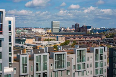 Modern şehir manzarası, önünde büyük pencereler olan beyaz önlü binalar, arkasında çeşitli mimari yapılar, yarı bulutlu bir gökyüzü altında muhtemelen Kopenhag.