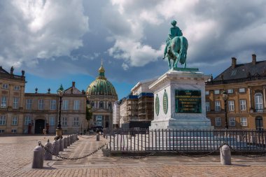 Tarihi bir Avrupa meydanında, muhtemelen Kopenhag 'da bulunan barok kubbeli klasik binalarla çevrili taş bir kaide üzerinde oksitlenmiş bronz bir binicilik heykeli yer alıyor..