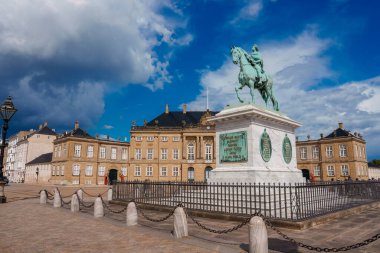 Kopenhag 'da parçalı bulutlu bir gökyüzünün altında, klasik sarı bir binanın yakınındaki kaldırımlı taşlı bir meydanda sergilenen, bir yamaç üzerinde orta dörtnala giden binaların heykeli..