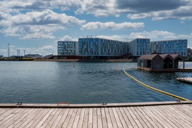 Kopenhag 'da ahşap bir iskelesi, metal kramponu ve turuncu yaşam halkası olan sakin bir rıhtım, modern binalar, sarı bir bariyer hattı ve parçalı bulutlu bir gökyüzü altında yüzen yapılar yer alıyor..