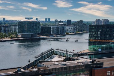 Kopenhag 'ın panoramik gündüz manzarası berrak gökyüzü, su, tekne ve rıhtım sakinlerini yansıtıyor. Çağdaş mimari ve devam eden inşaat kentsel büyümeyi vurguluyor.